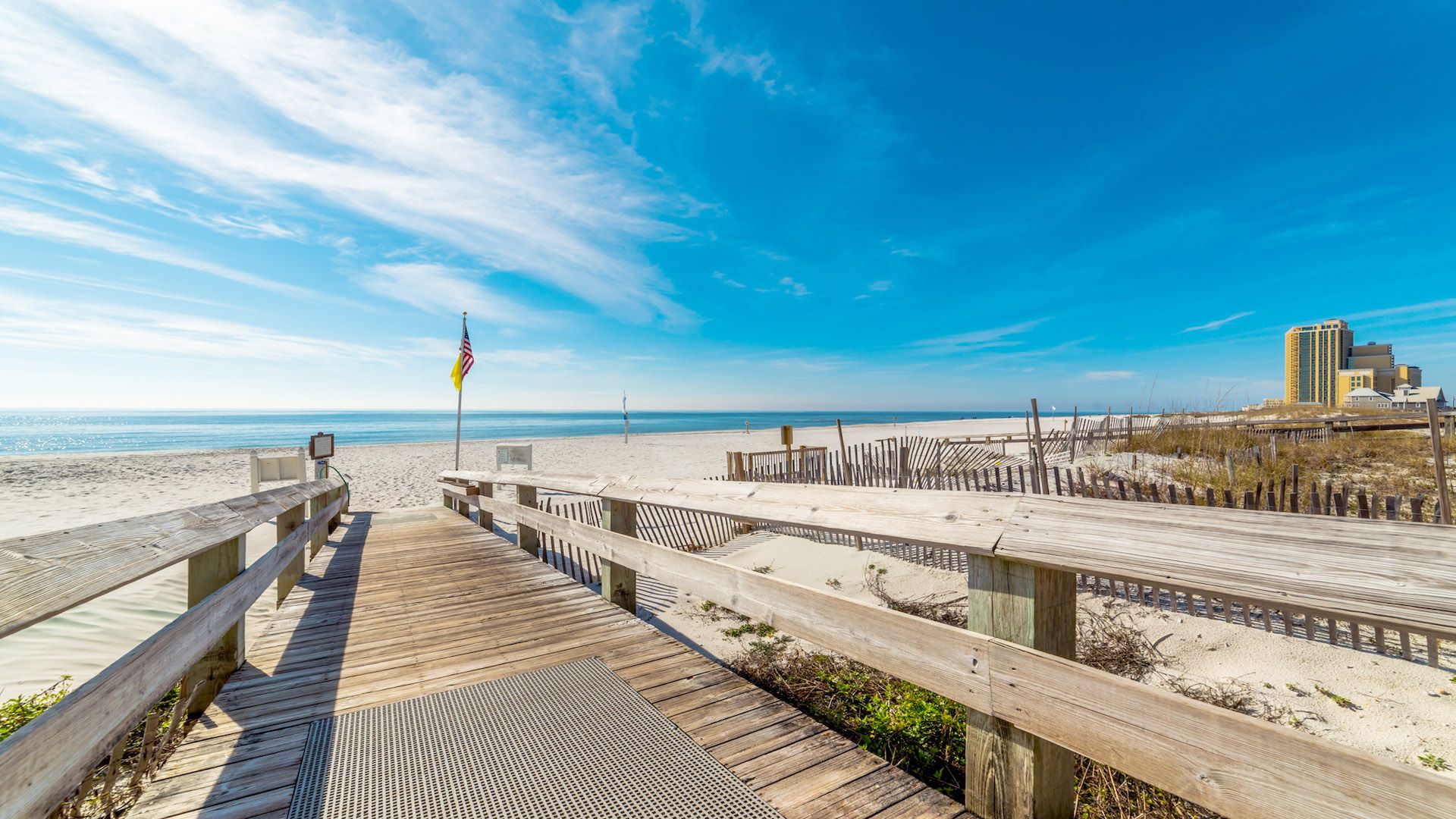 A wooden boardwalk leading to a sandy beach with a flag, a clear sky, and a building in the background.