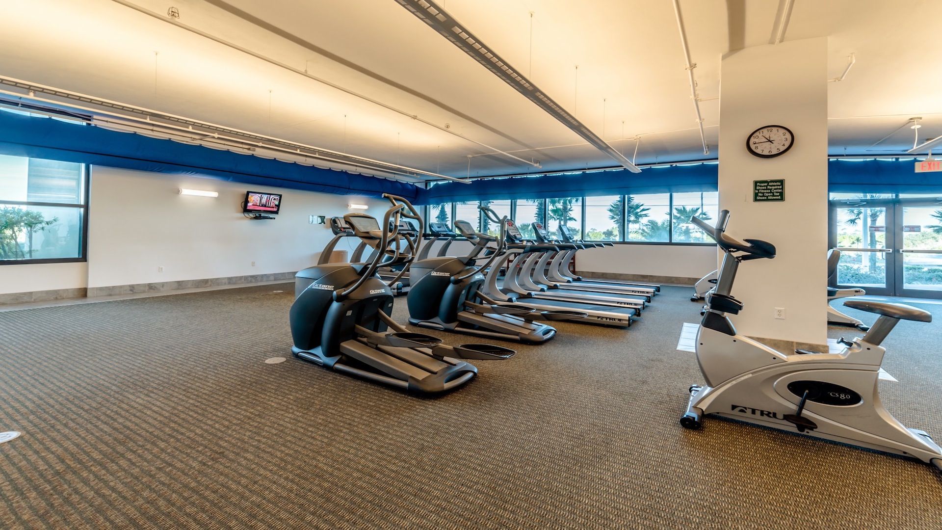 A gym with multiple treadmills and elliptical machines, large windows, and a clock on the wall.