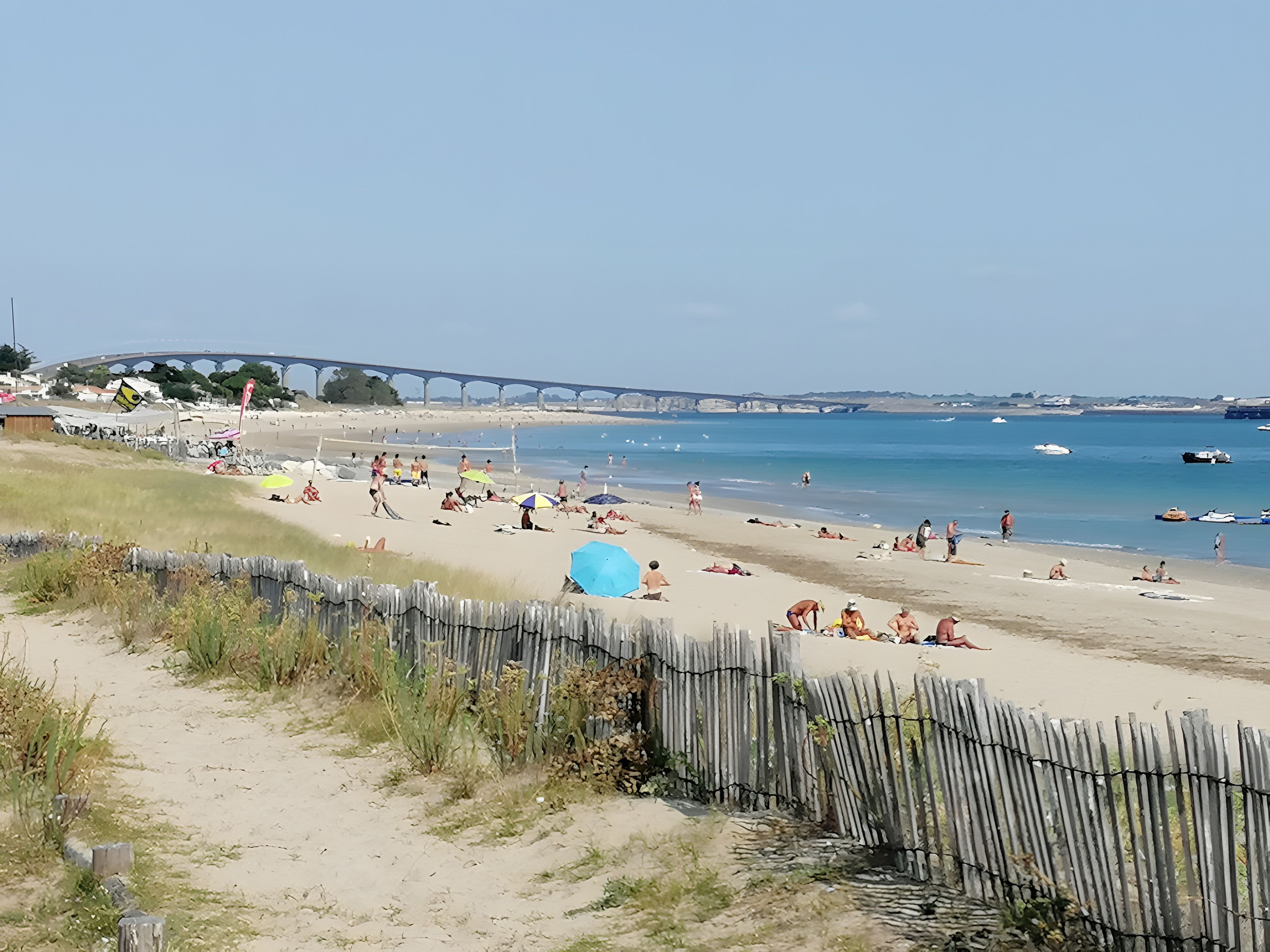 Une plage avec une clôture en bois, des gens qui prennent un bain de soleil et un pont en arrière-plan.