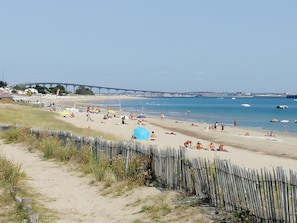 Ein Strand mit einer Holzbank, Menschen, die sich sonnen, und einer Brücke im Hintergrund.