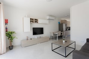 A modern living room with a flat-screen TV, a dining area, and a kitchen in the background.