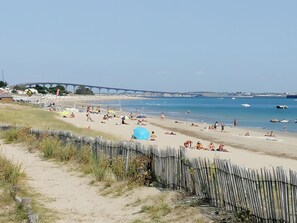 Ein Strand mit einer Holzbank, Menschen, die sich sonnen, und einer Brücke im Hintergrund.