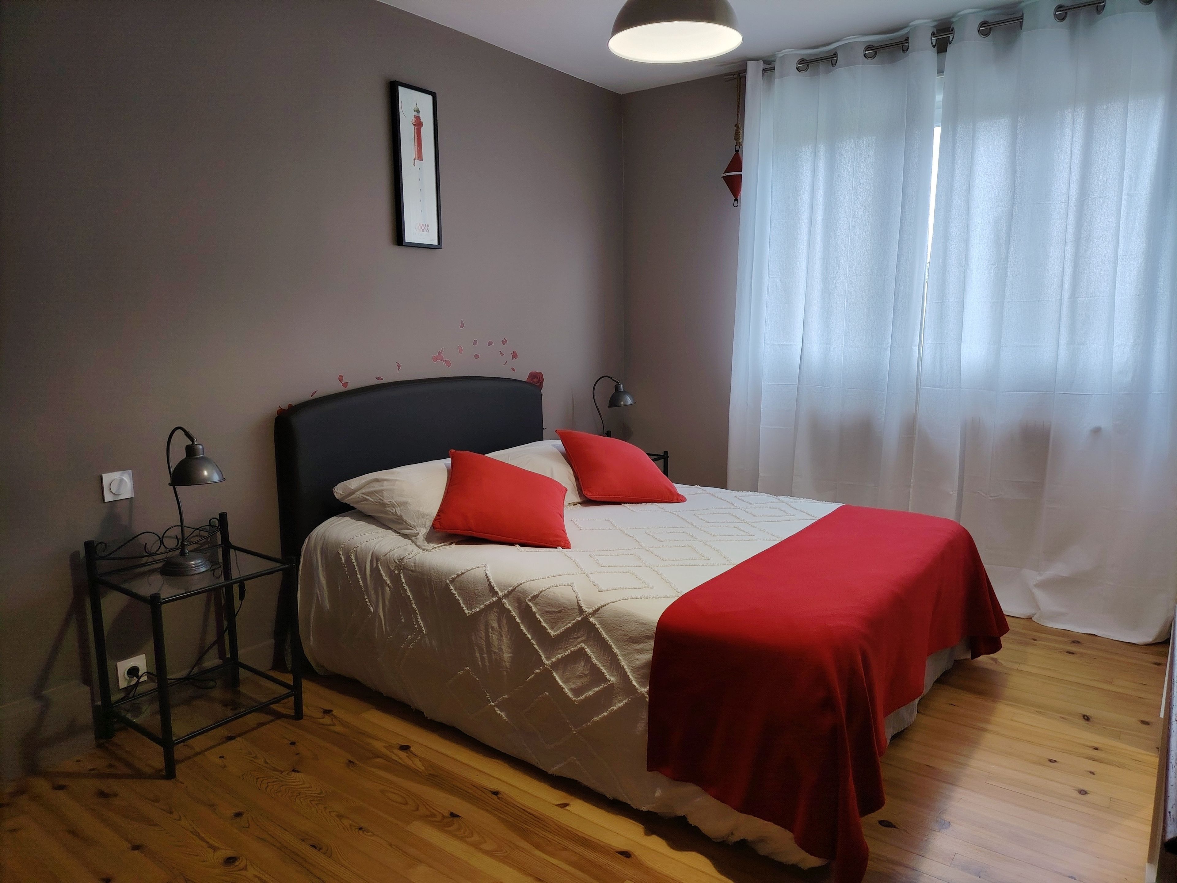 Un dormitorio con cama, mesita de noche, lámpara, un cuadro enmarcado en la pared y ventana con cortinas.