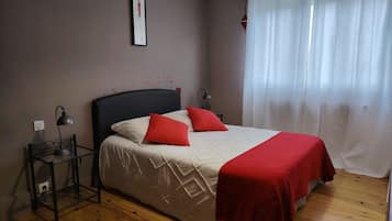 Un dormitorio con cama, mesita de noche, lámpara, un cuadro enmarcado en la pared y ventana con cortinas.