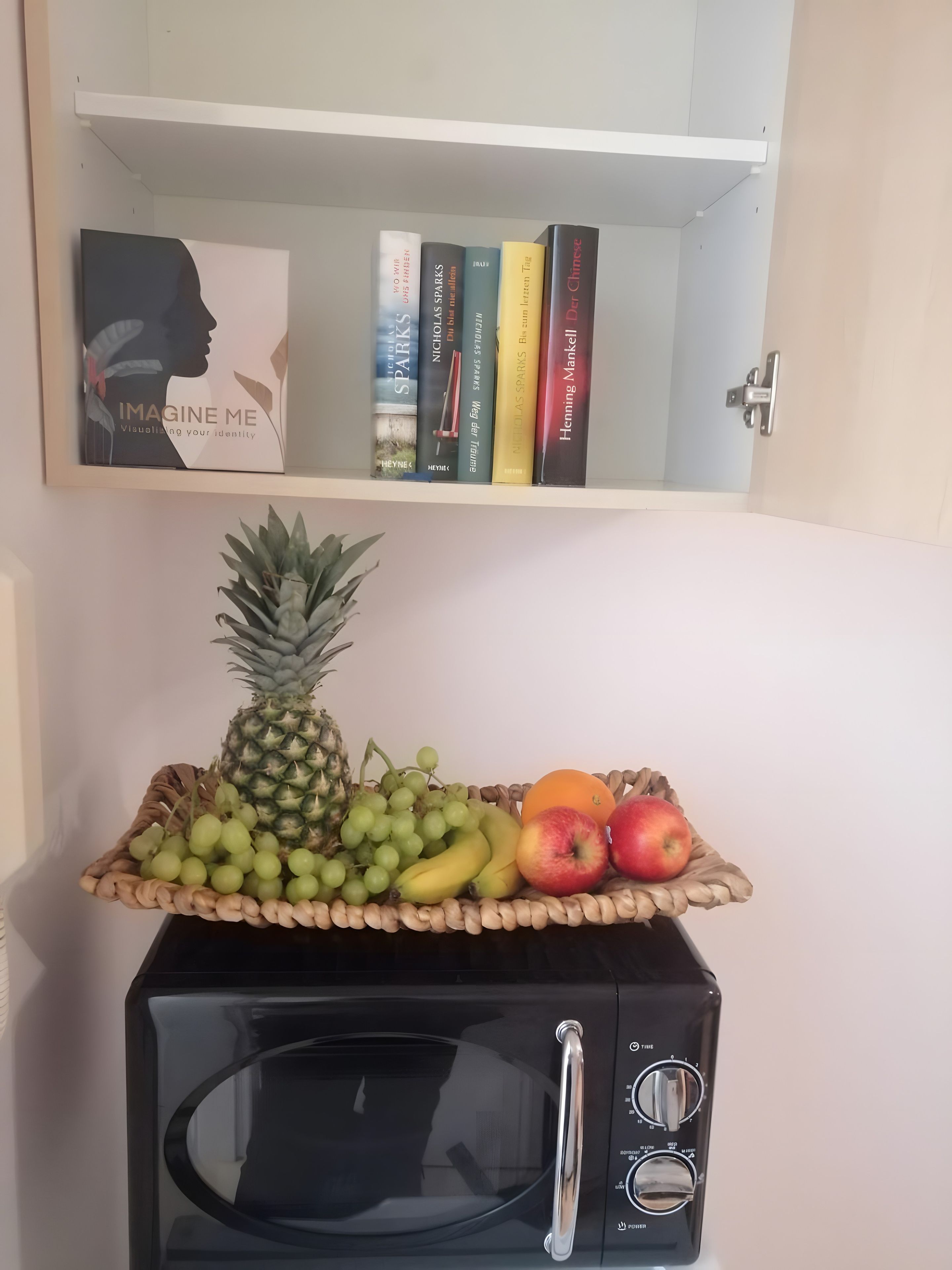 Una cocina con microondas, una cesta de fruta encima y una estantería con libros.
