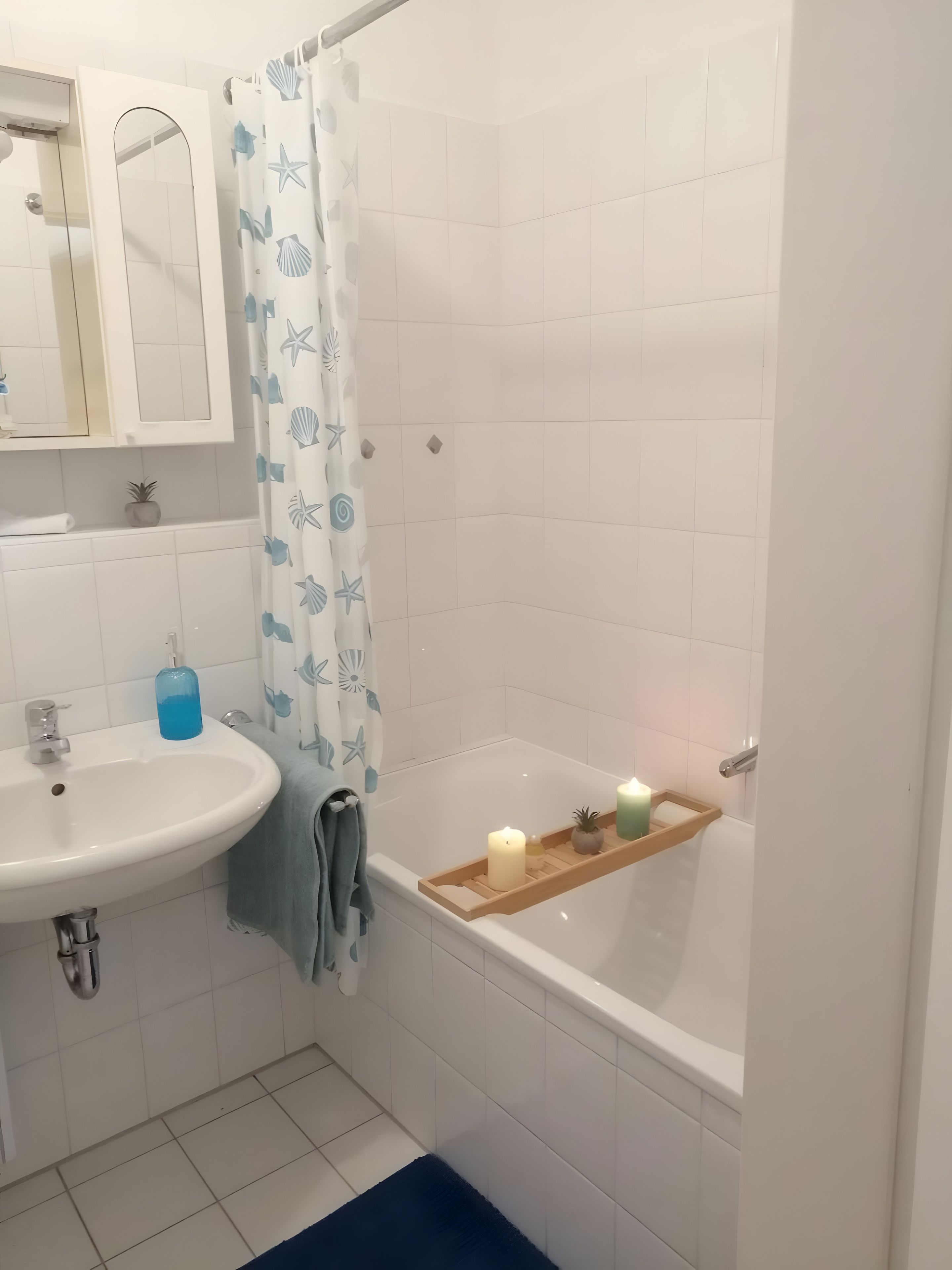 Ein Badezimmer mit einem weißen Waschbecken, Spiegel und Badewanne. Ein blaues Handtuch und ein Ablagefach mit Kerzen sind vorhanden.