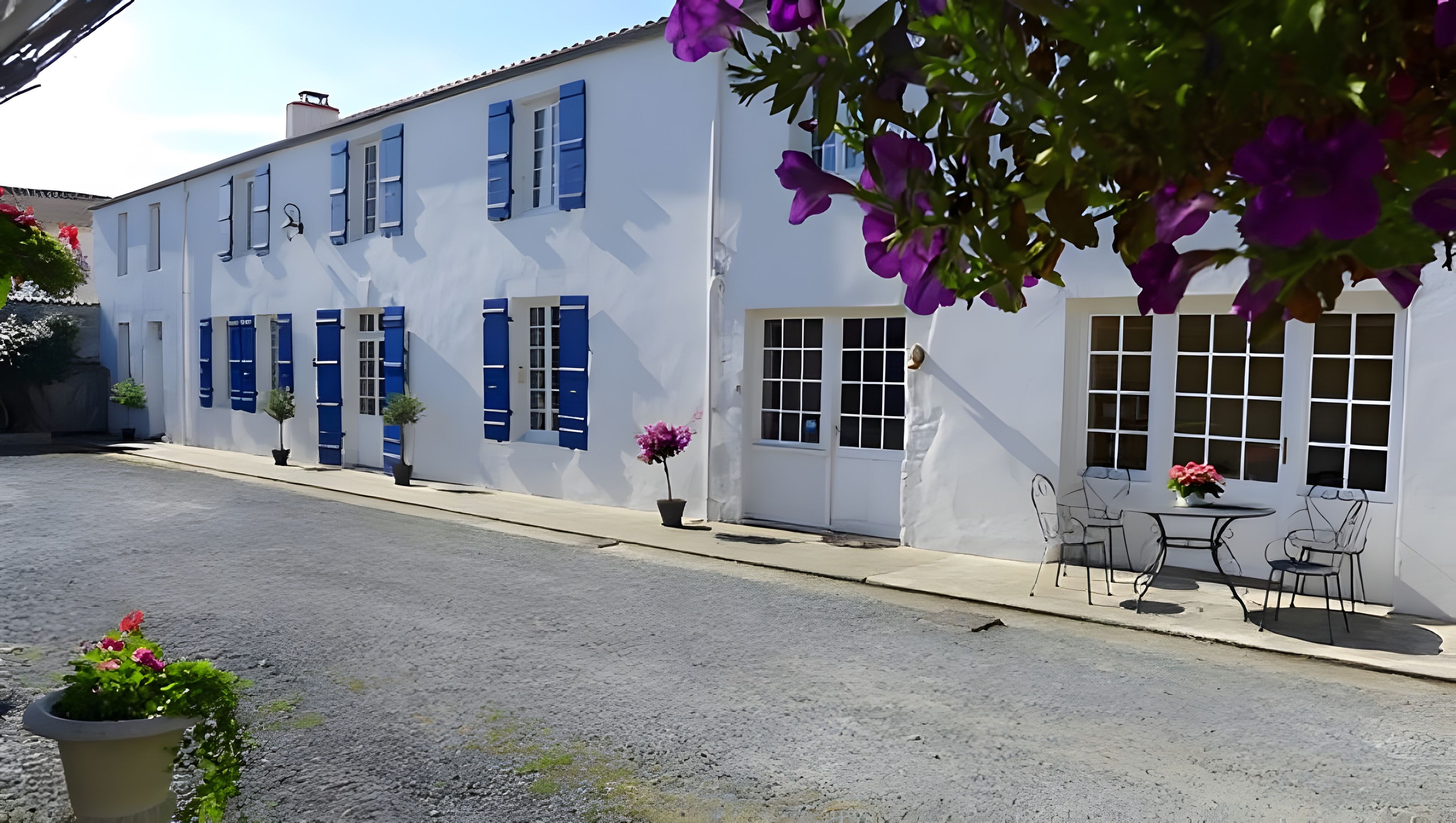A white building with blue shutters, a door, and windows. There is a small outdoor table with chairs and a potted plant.