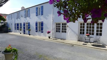 A white building with blue shutters, a door, and windows. There is a small outdoor table with chairs and a potted plant.
