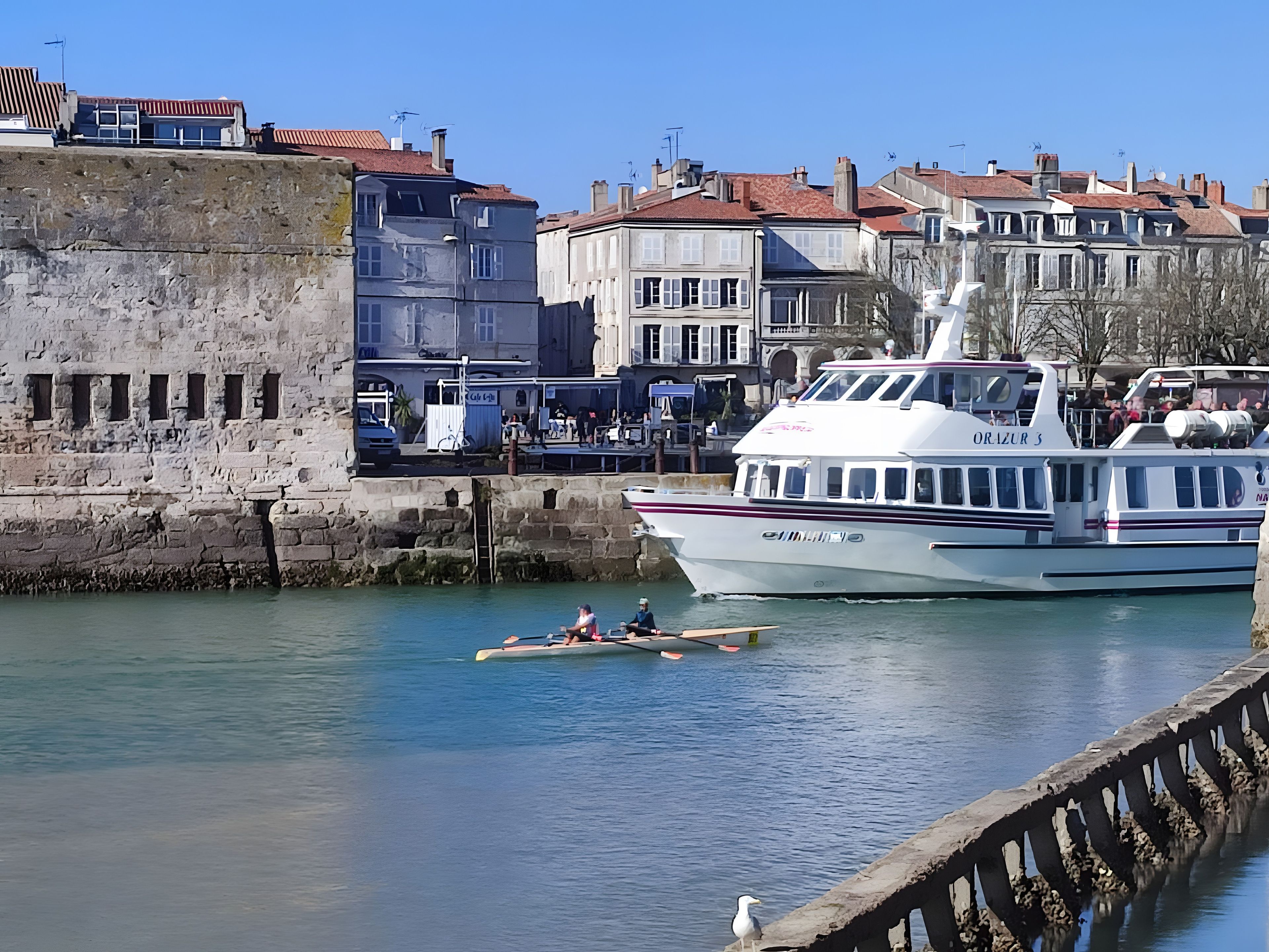 Une scène de bord de mer avec un grand bateau blanc amarré à un quai, un mur de pierre et, au fond, des bâtiments aux toits rouges.