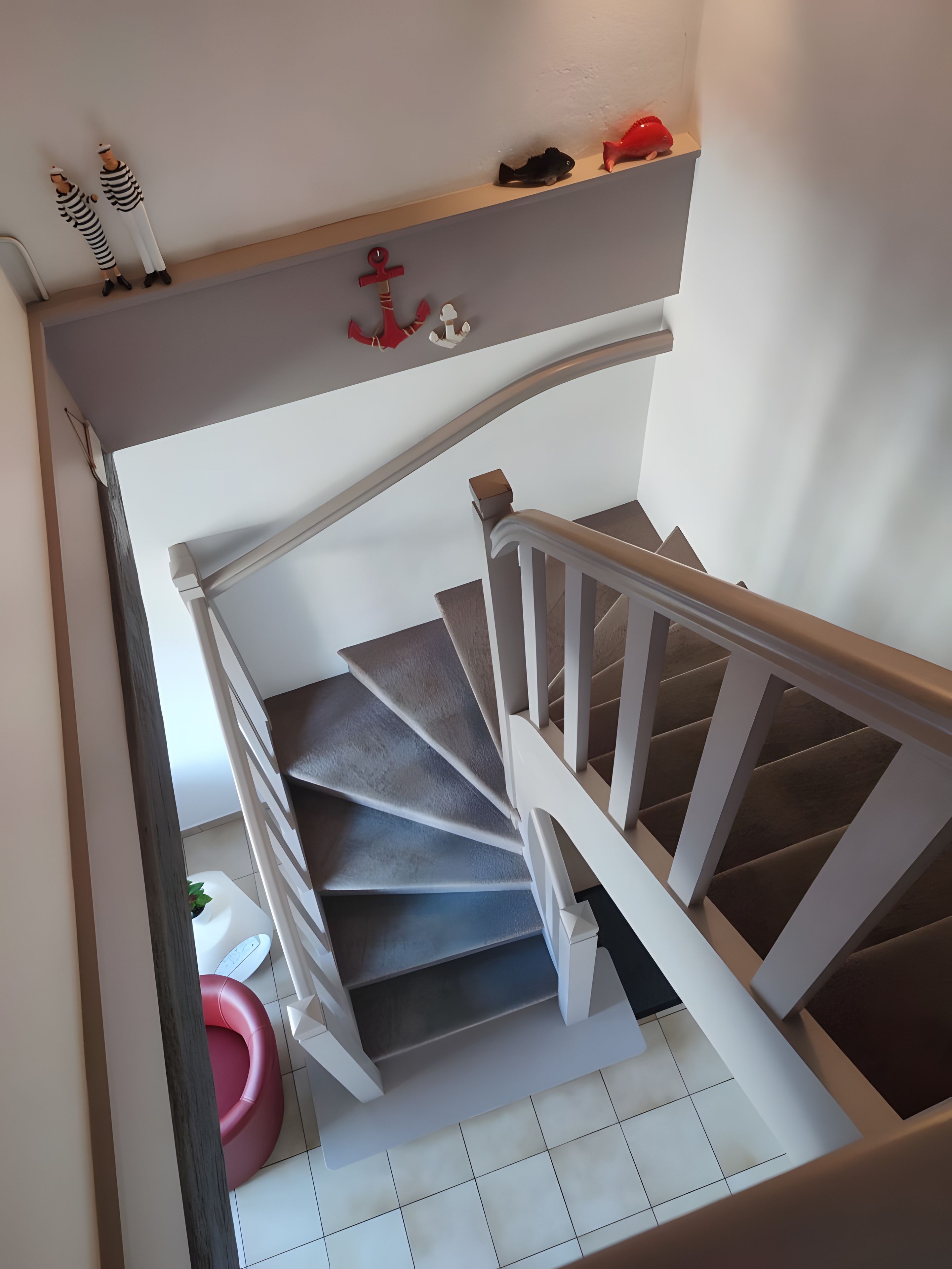 Un escalier avec une rampe blanche et des marches grises, menant à un palier où se trouve une petite étagère sur laquelle sont disposés des objets décoratifs.