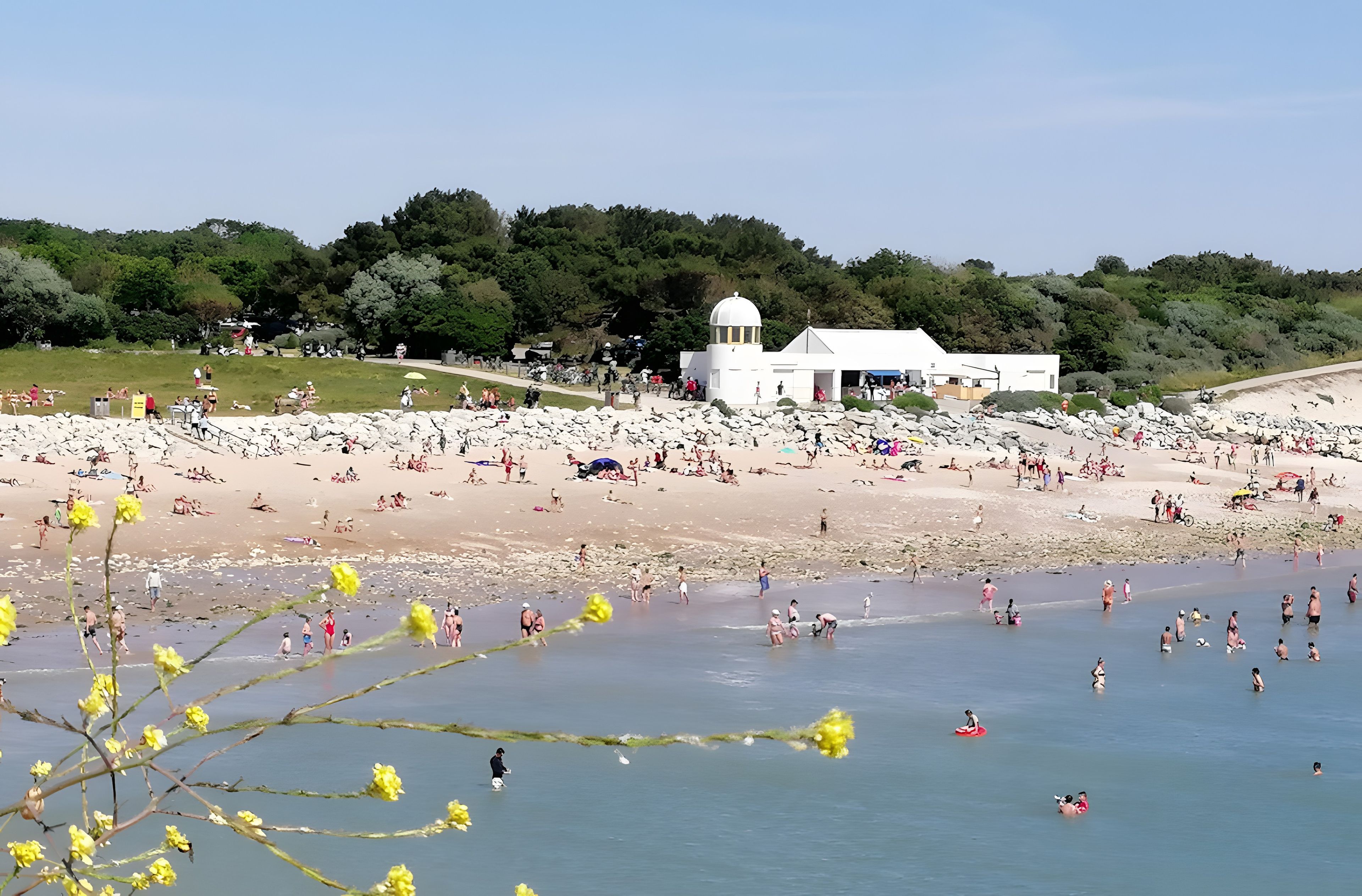 Une plage avec un bâtiment blanc, des gens qui prennent un bain de soleil et un ciel dégagé.