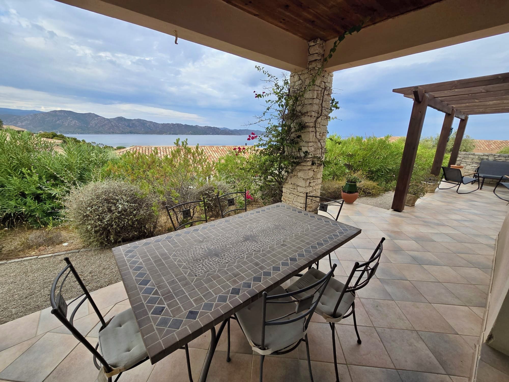Eine Terrasse im Freien mit Tisch und Stühlen, mit Blick auf ein Gewässer und Berge.