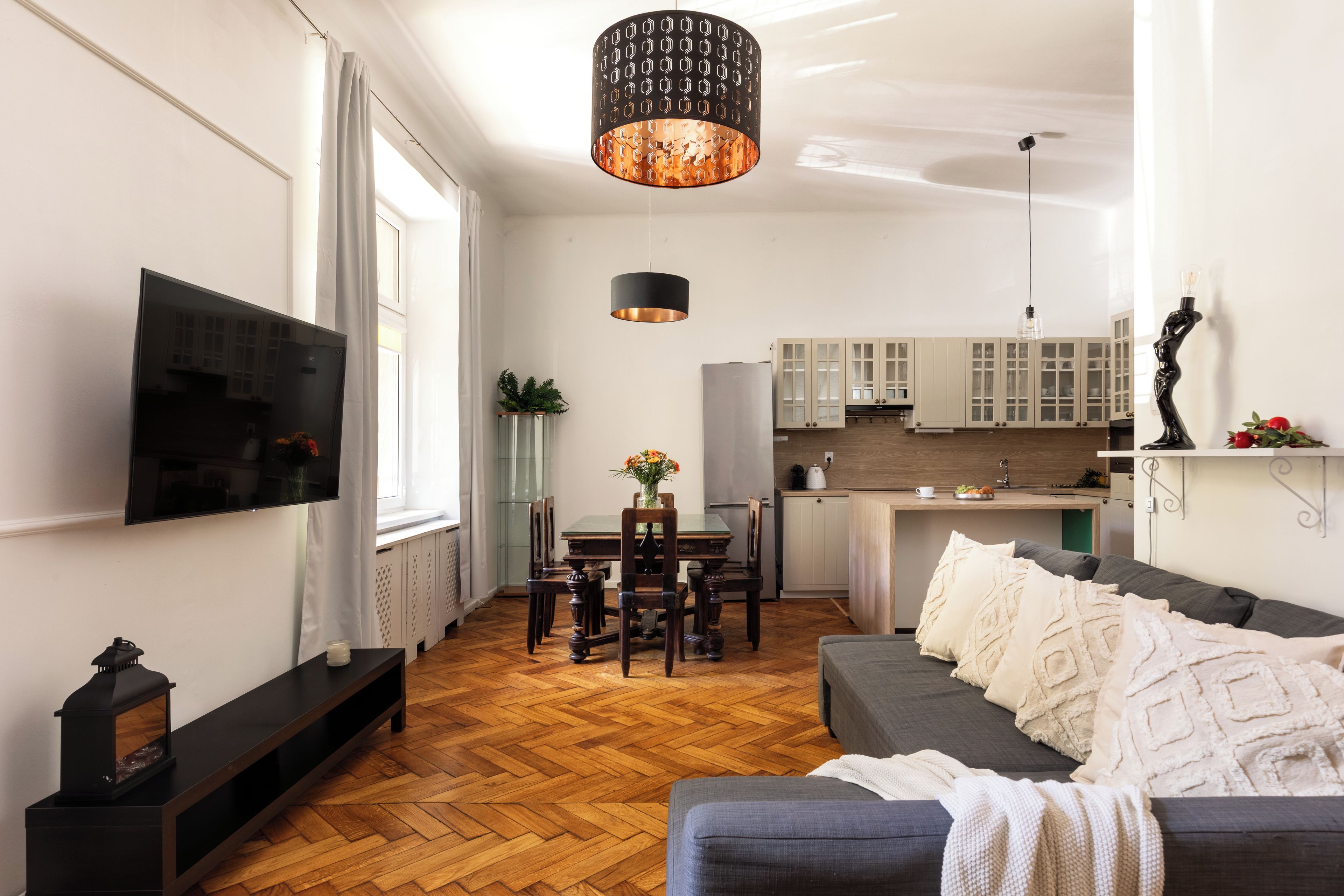 A modern living room with a flat-screen TV, a sofa, a dining table with chairs, a kitchen area with cabinets, and a hanging pendant light.