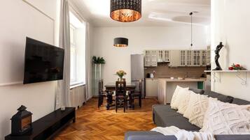 A modern living room with a flat-screen TV, a sofa, a dining table with chairs, a kitchen area with cabinets, and a hanging pendant light.