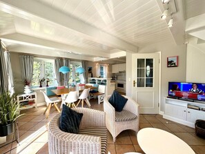 A spacious living area with a dining table, wicker chairs, a television, and a kitchen in the background. - Westerhus Nieblum (Nieblum)