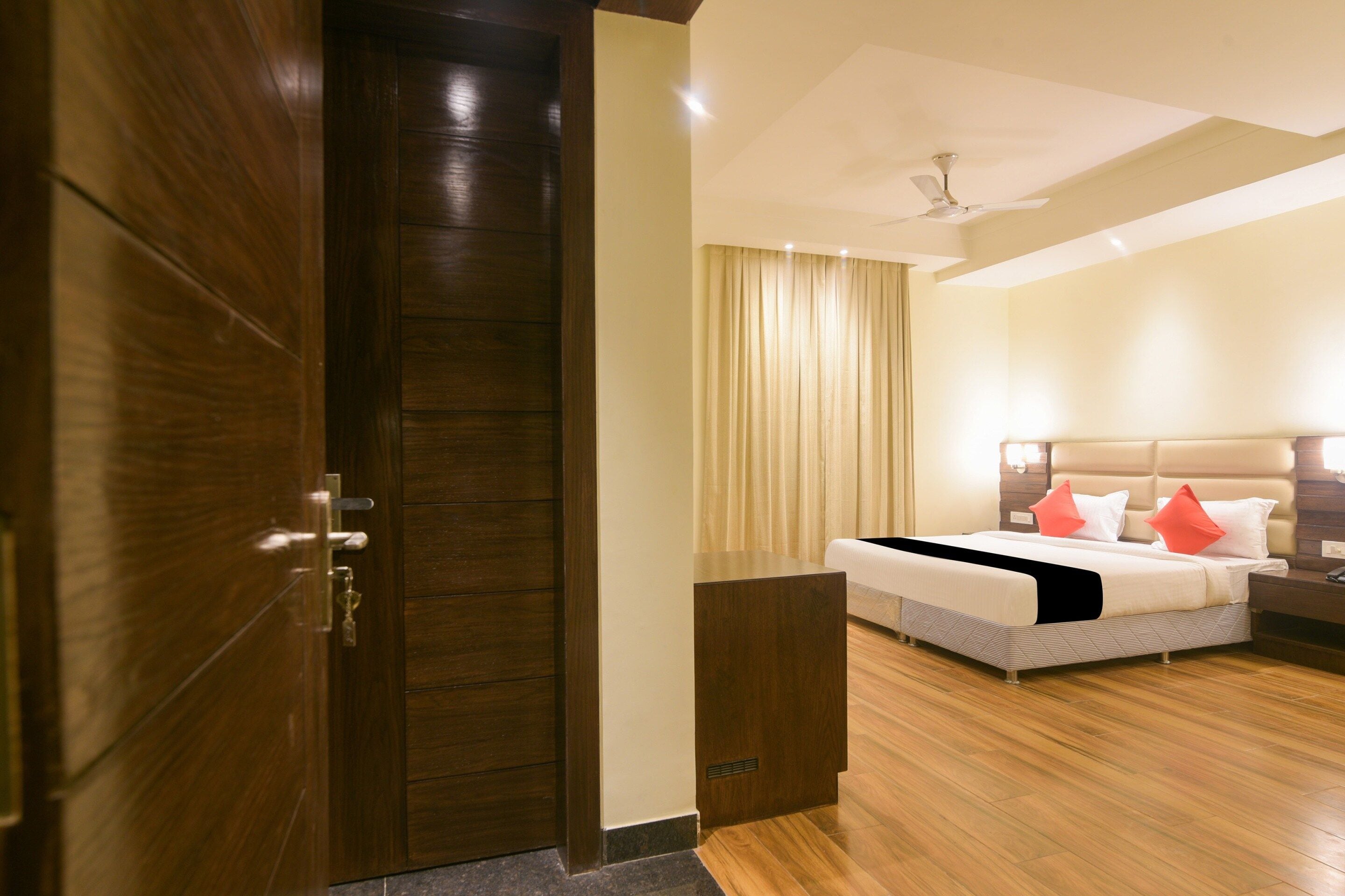 A hotel room with a wooden door, a bed with white bedding and red pillows, a nightstand, and a ceiling fan.