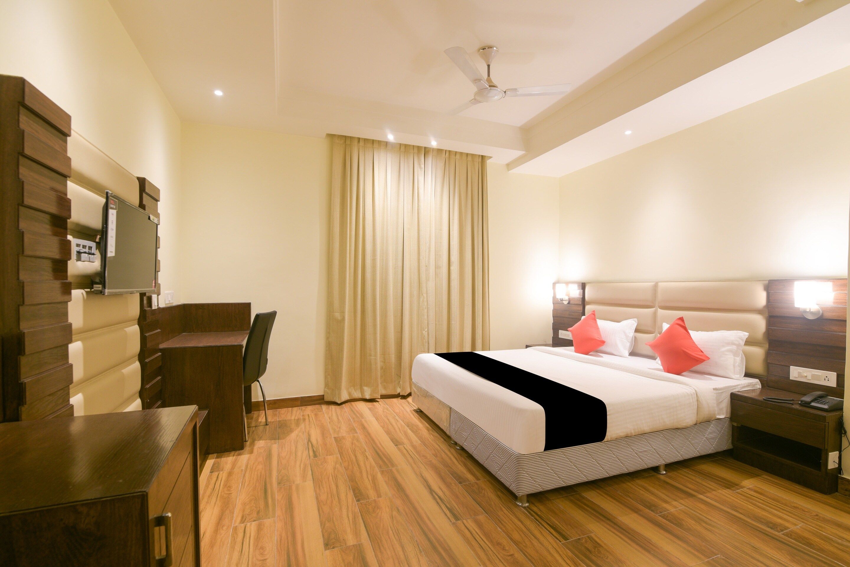 Quarto de hotel com cama, mesa de cabeceira, cadeira, escrivaninha e ventilador de teto.