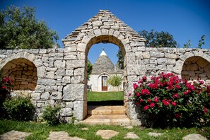 Un arc en pierre mène à un bâtiment surmonté d’un toit conique, le tout étant entouré de verdure et de fleurs.
