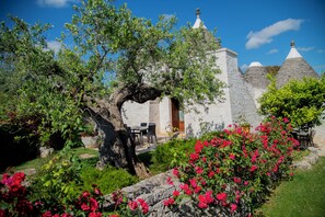 Un bâtiment en pierre, doté d’une façade blanche et de toits coniques, entouré d’une végétation luxuriante et d’arbustes en fleurs.