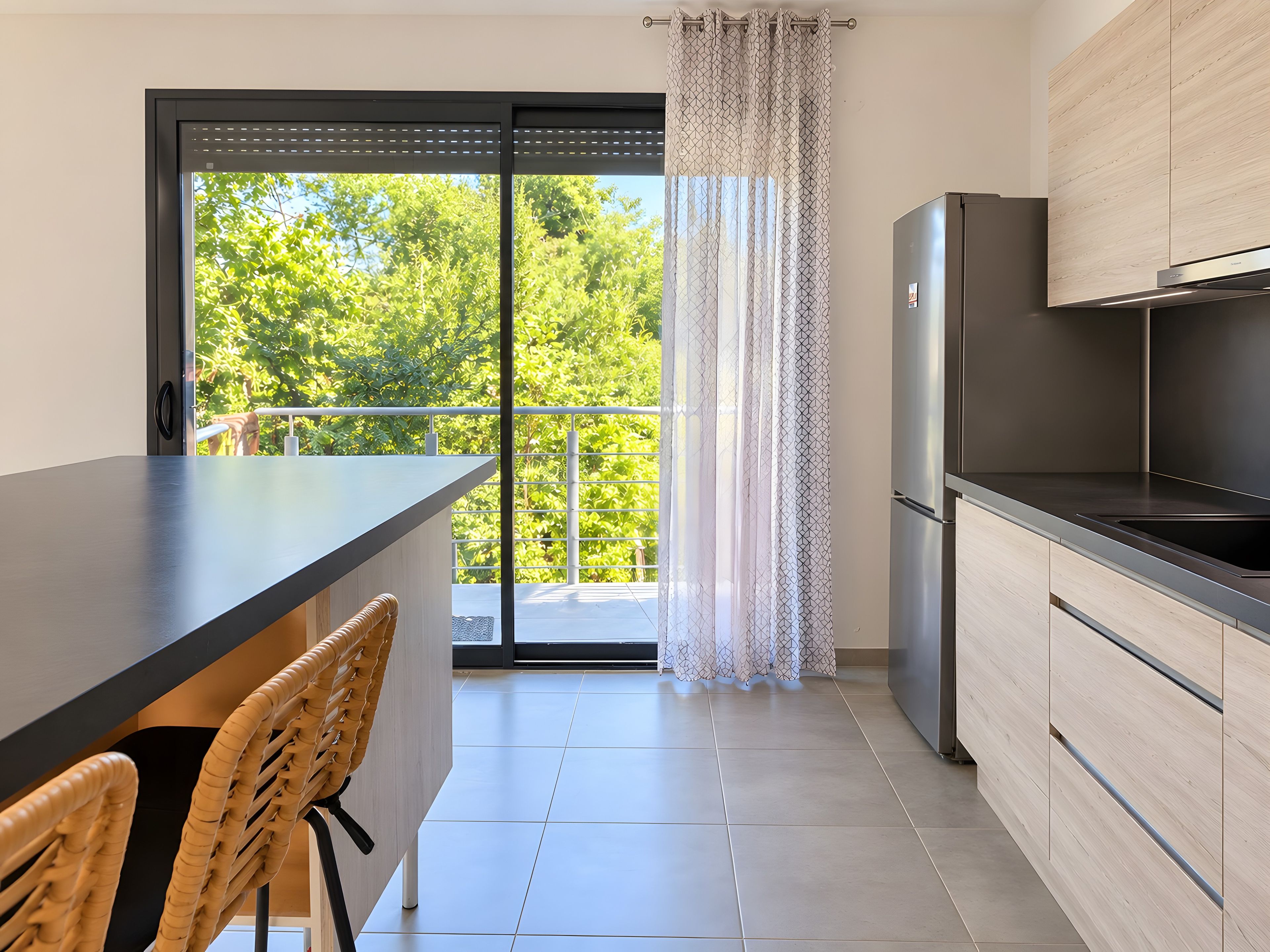 Una cocina moderna con una isla grande, un refrigerador y una puerta corrediza de vidrio que da a un balcón con vistas a árboles.