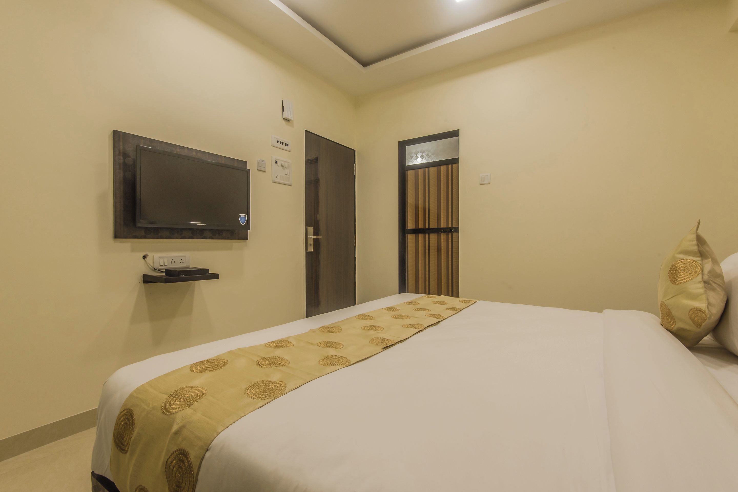 Habitació d'hotel amb un llit, una televisió penjada a la paret, una porta i una petita estant amb un aparell.