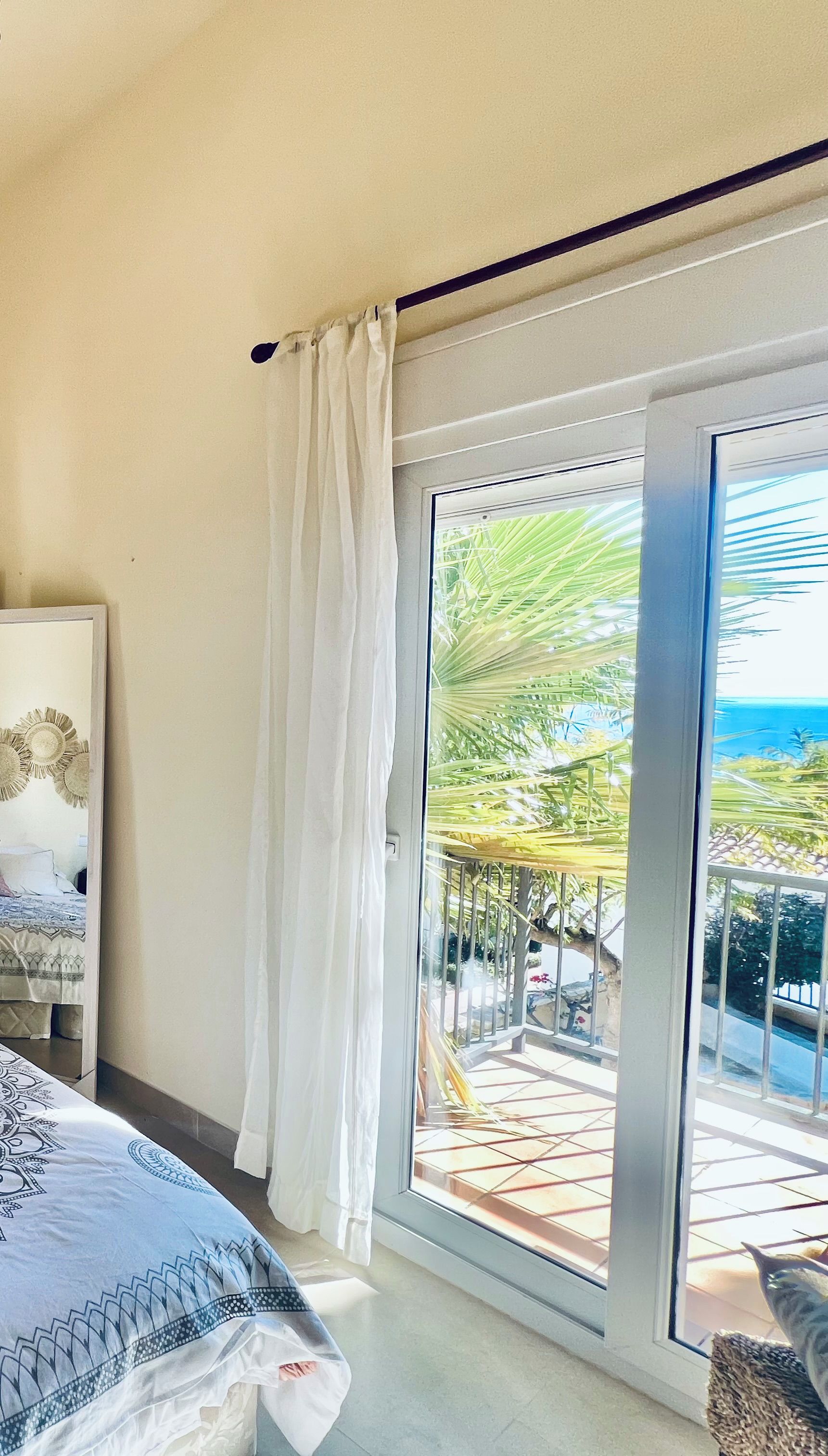 A bedroom with a bed, a window with curtains, and a view of a balcony with outdoor furniture.