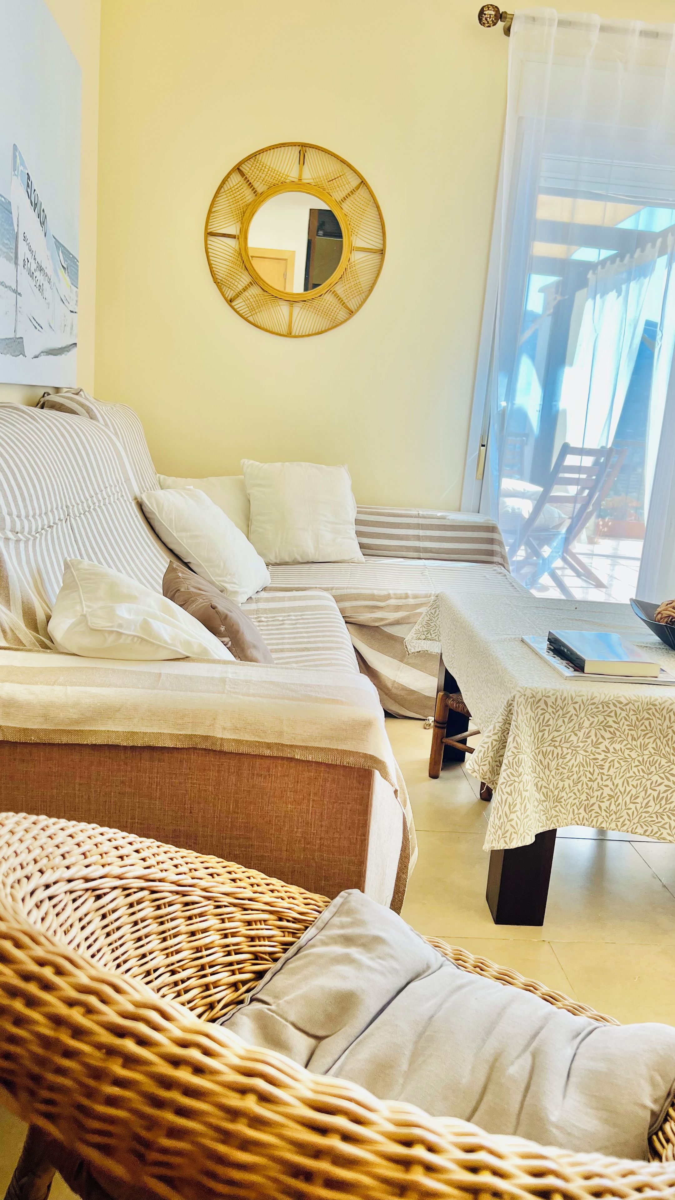 A room with a sofa set, a wicker chair, a small table with a book, and a round mirror on the wall.