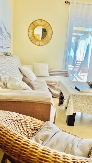 A room with a sofa set, a wicker chair, a small table with a book, and a round mirror on the wall. - Vrbo Property (AN)