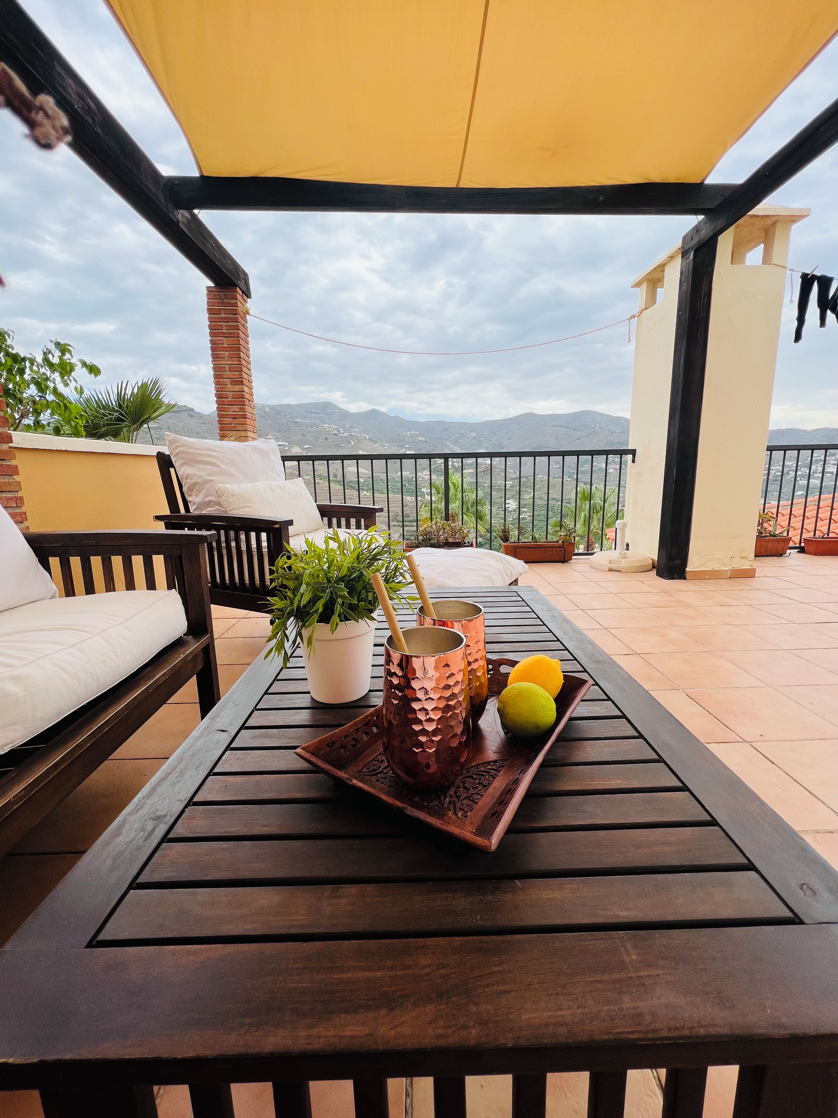 A wooden table with a tray holding two copper mugs and lemons, a potted plant, and a white cushion on a balcony with a yellow canopy, overlooking a mountainous landscape.