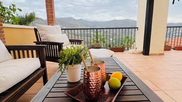 A wooden table with a tray holding two copper mugs and lemons, a potted plant, and a white cushion on a balcony with a yellow canopy, overlooking a mountainous landscape.
