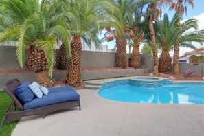 A swimming pool with a curved edge, surrounded by palm trees and a lounge chair with blue cushions.