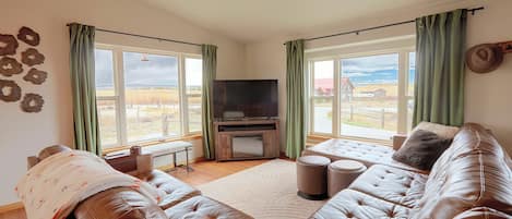 A living room with a leather sofa, a flat-screen TV, and large windows with curtains.