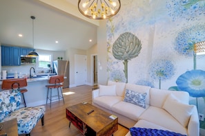 Ein modernes Wohnzimmer mit einer Couch, einem Couchtisch und Sesseln. Es gibt eine Küchenzeile mit blauen Schränken und einem Esstisch. An der Wand befindet sich eine große Blumenwandmalerei.