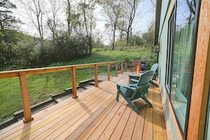Eine Holzterrasse mit Glastrennwand, Sitzmöbeln und Blick auf eine grüne Landschaft.