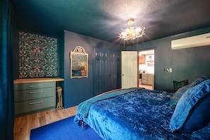 Un dormitorio con cama, cómoda, una puerta y una pequeña cocina visible a través de una puerta abierta.
