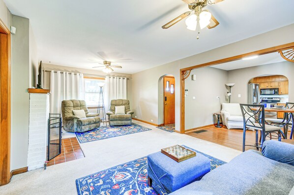A spacious living room with a ceiling fan, two armchairs, a coffee table, and a kitchen area with a microwave and refrigerator.