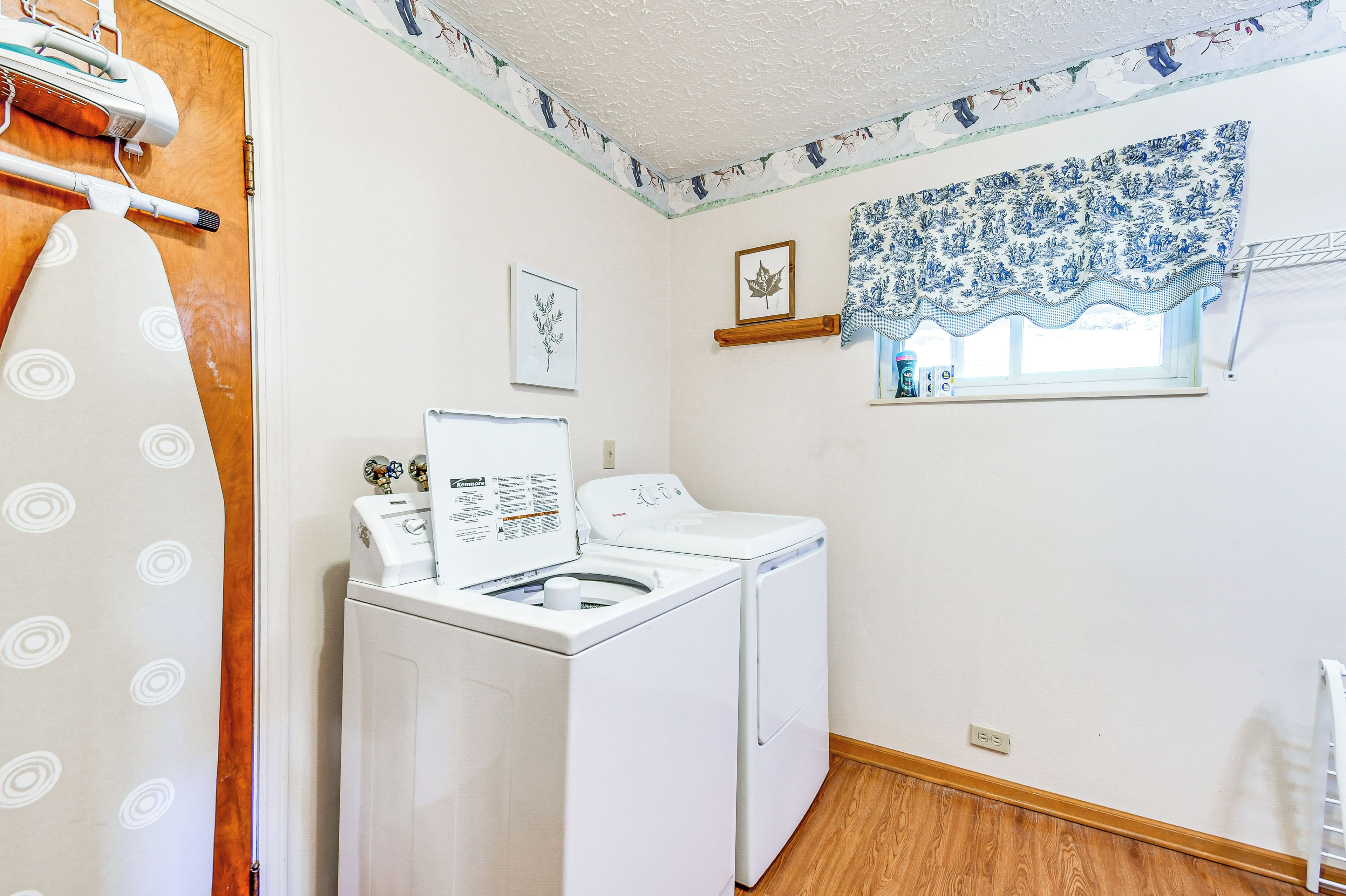 A laundry room with a Kenmore washer and dryer, a wooden door, a window with blue floral curtains, and a wall-mounted shelf with a framed leaf picture.