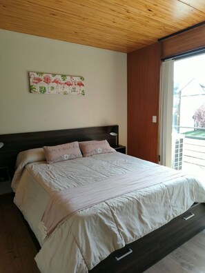 A bedroom with a bed, wooden ceiling, a wall with a floral painting, and a window with a view of buildings. - Apartment "Plaza Sarmiento" I—Centrally Located with a Beautiful View (San Martín de los Andes)