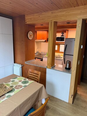 Cocina compacta con armarios de madera, microondas, refrigerador y comedor con mesa y sillas.