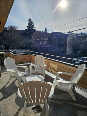 A balcony with white plastic chairs and a small round table, overlooking a street with buildings and parked cars. - Apartment "Plaza Sarmiento" I—Centrally Located with a Beautiful View (San Martín de los Andes)