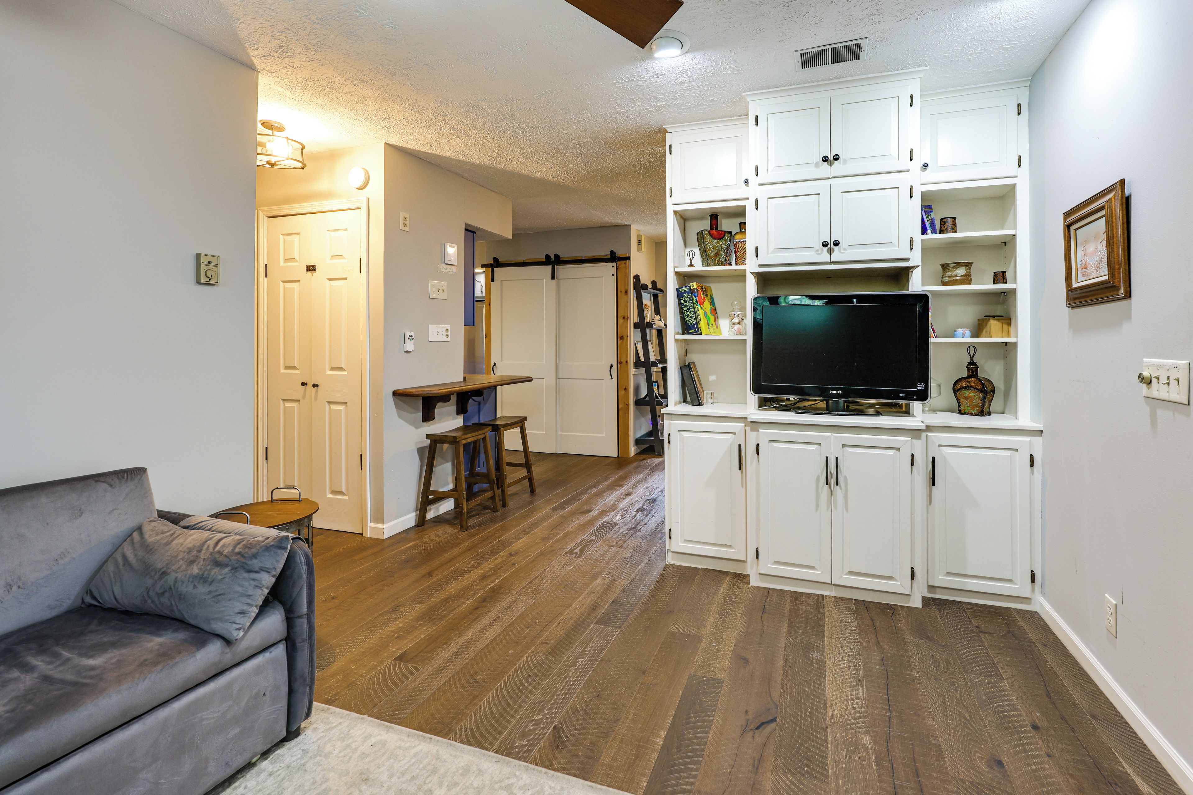 A living room with a grey sofa, wooden flooring, white cabinets, and a flat-screen TV.