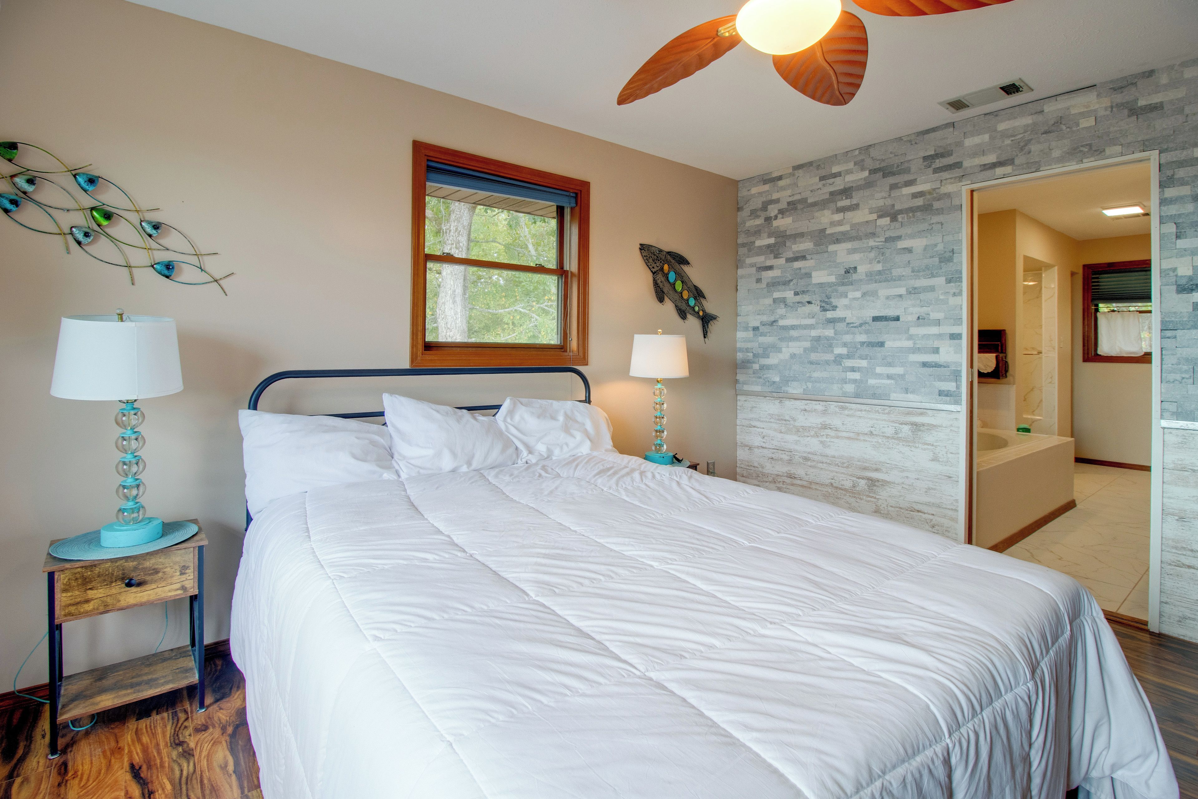 A bedroom with a bed, two bedside tables with lamps, a ceiling fan, a window with a view of trees, and a bathroom visible through an open door.