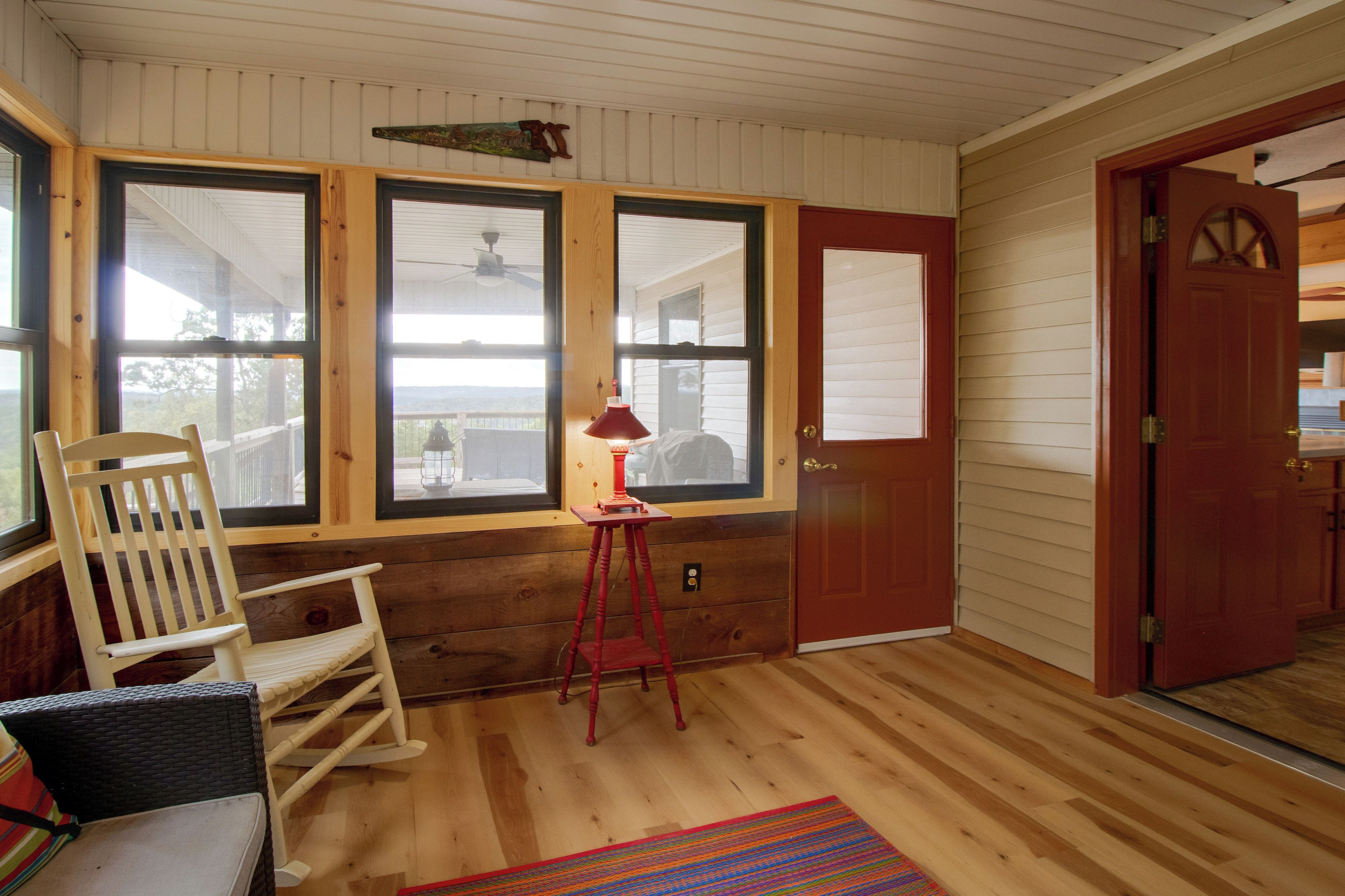 A room with wooden flooring, a rocking chair, a red lamp, and large windows.