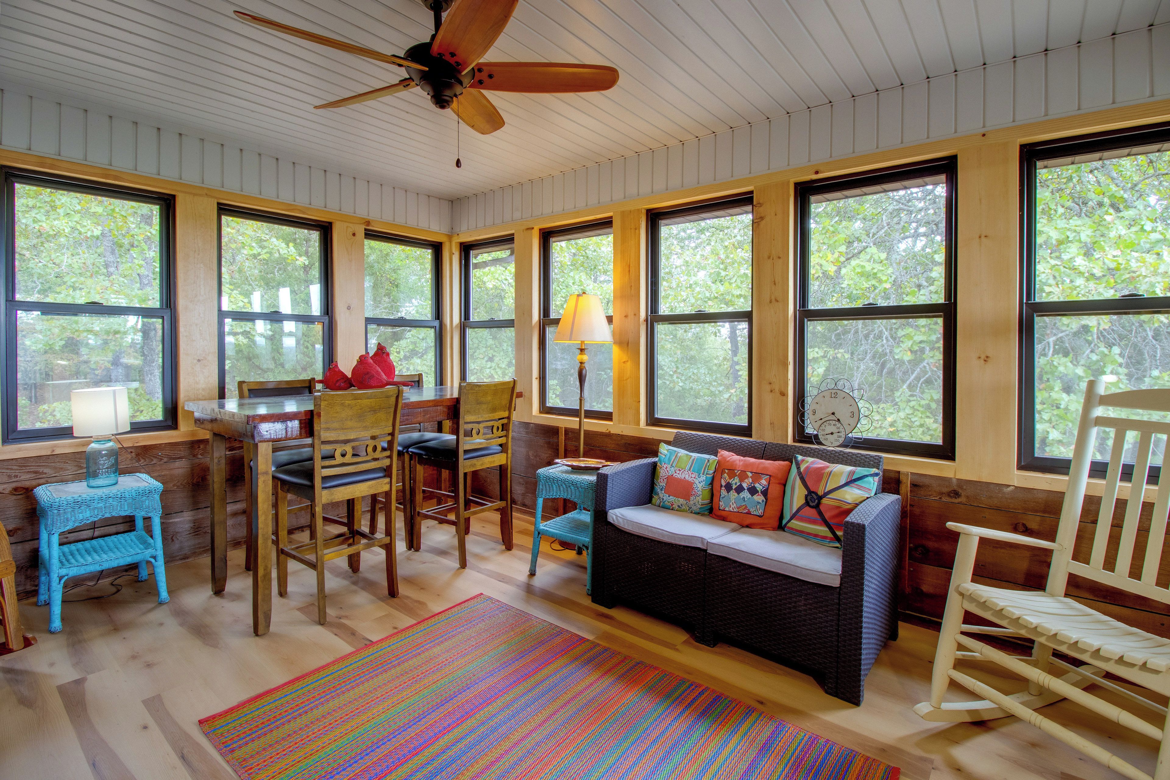 A room with a wooden ceiling fan, large windows, a dining table with chairs, a sofa with cushions, and a colorful rug.