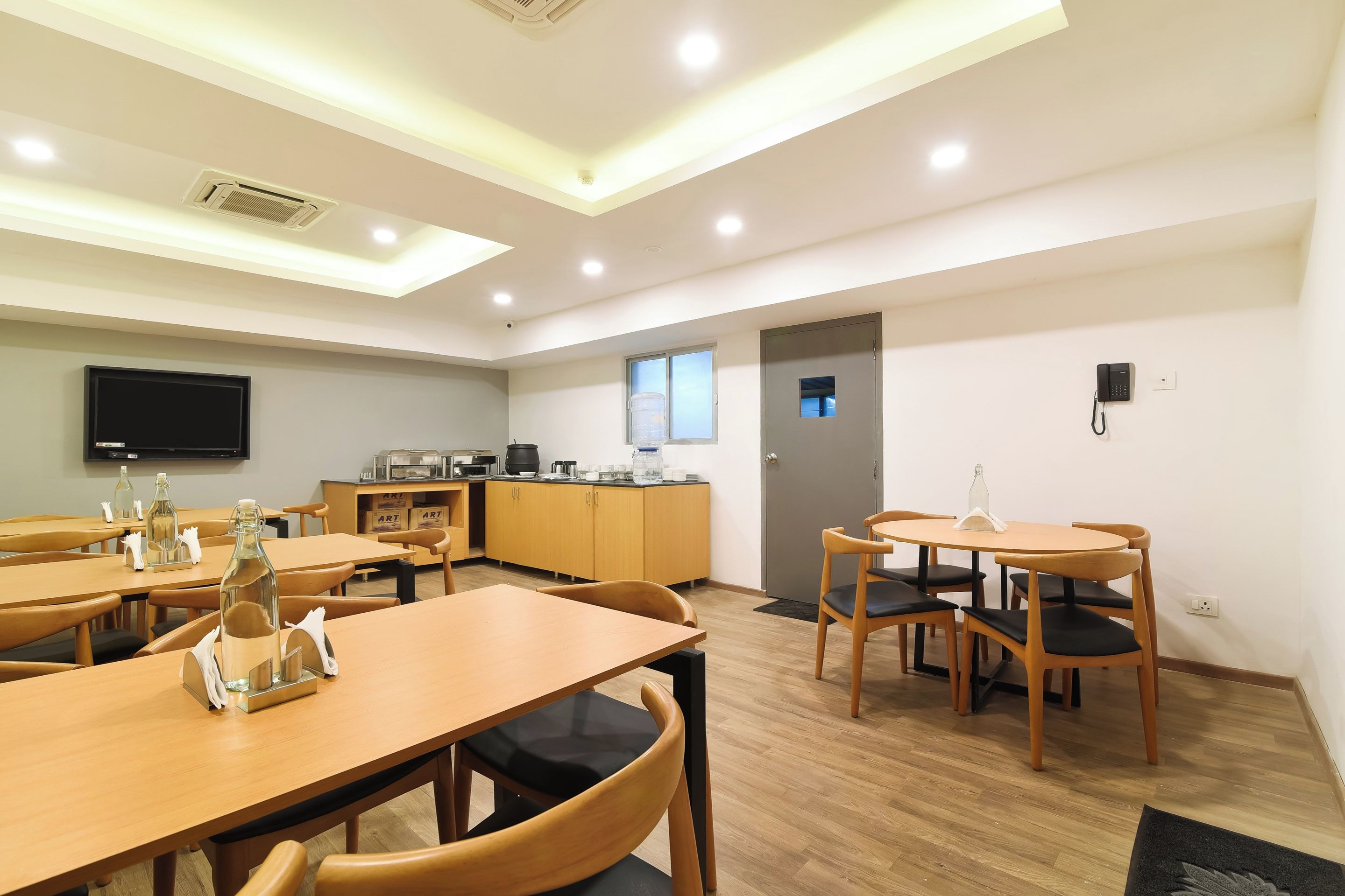 A hotel dining area with multiple wooden tables and chairs, a kitchen area with appliances, and a television mounted on the wall.