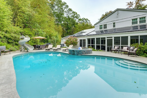 A swimming pool with lounge chairs and a slide, surrounded by trees and a building with large windows.