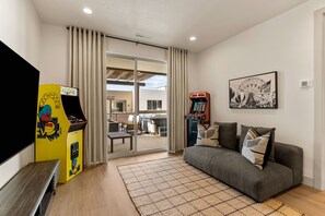 A living room with a grey sofa, a flat-screen TV, a Pac-Man arcade game, and a Ferris wheel artwork on the wall. - Six Keys: Personal Golf Cart + Hot Tub! (St. George)