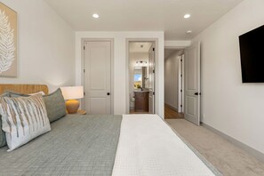 Une chambre à coucher comprenant un lit, une table de chevet, une lampe, un téléviseur fixé au mur et une porte donnant sur une salle de bain.