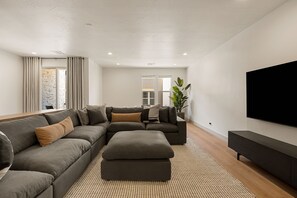 A modern living room with a sectional sofa, a coffee table, a flat-screen TV, and a large window with curtains. - Six Keys: Personal Golf Cart + Hot Tub! (St. George)