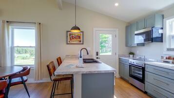 A modern kitchen with a central island, bar stools, a dining table, and a view of the outdoors through a door.